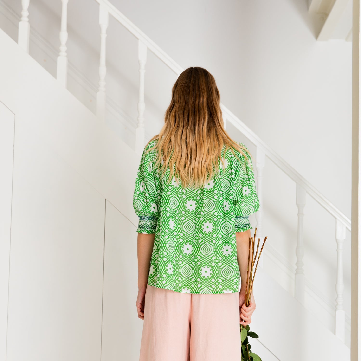 Woman from behind wearing a green, geometric-patterned blouse and pink pants, holding stems of flowers.