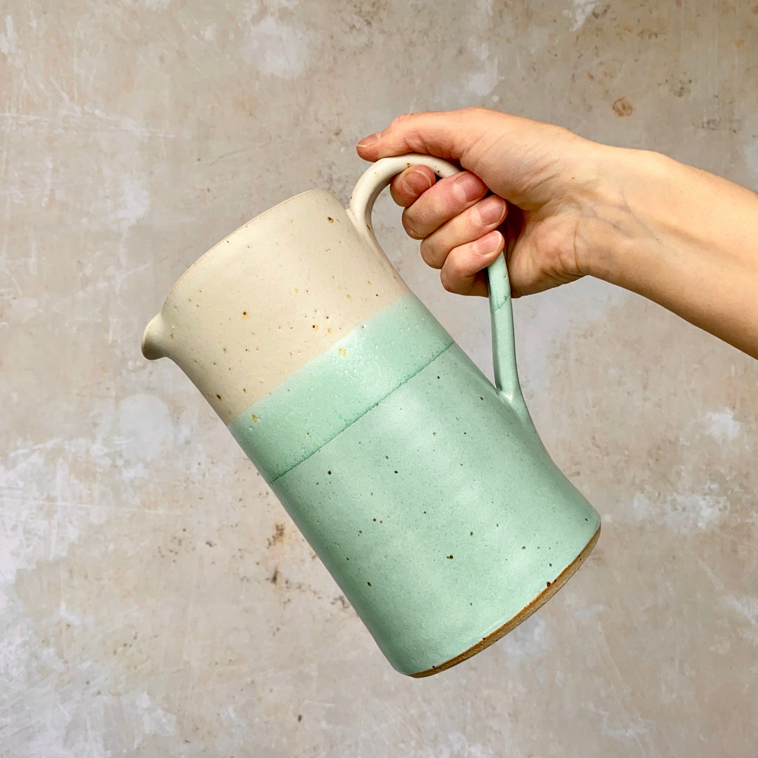 Hand holding a speckled, two-toned ceramic pitcher.  The top half is off-white, the bottom a pale seafoam green.