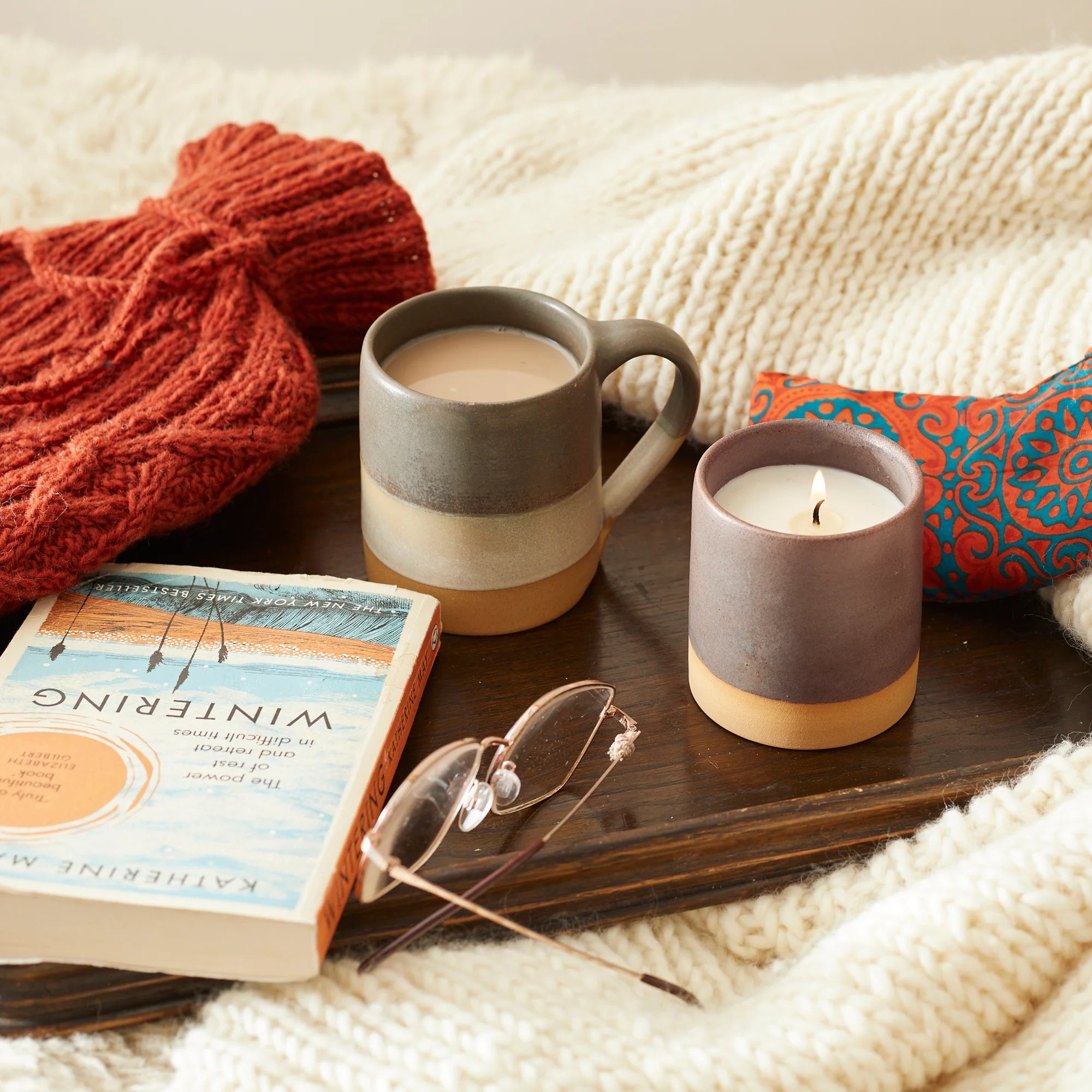 A lit candle in a dark gray and tan ceramic jar, next to a mug of tea on a wooden tray.