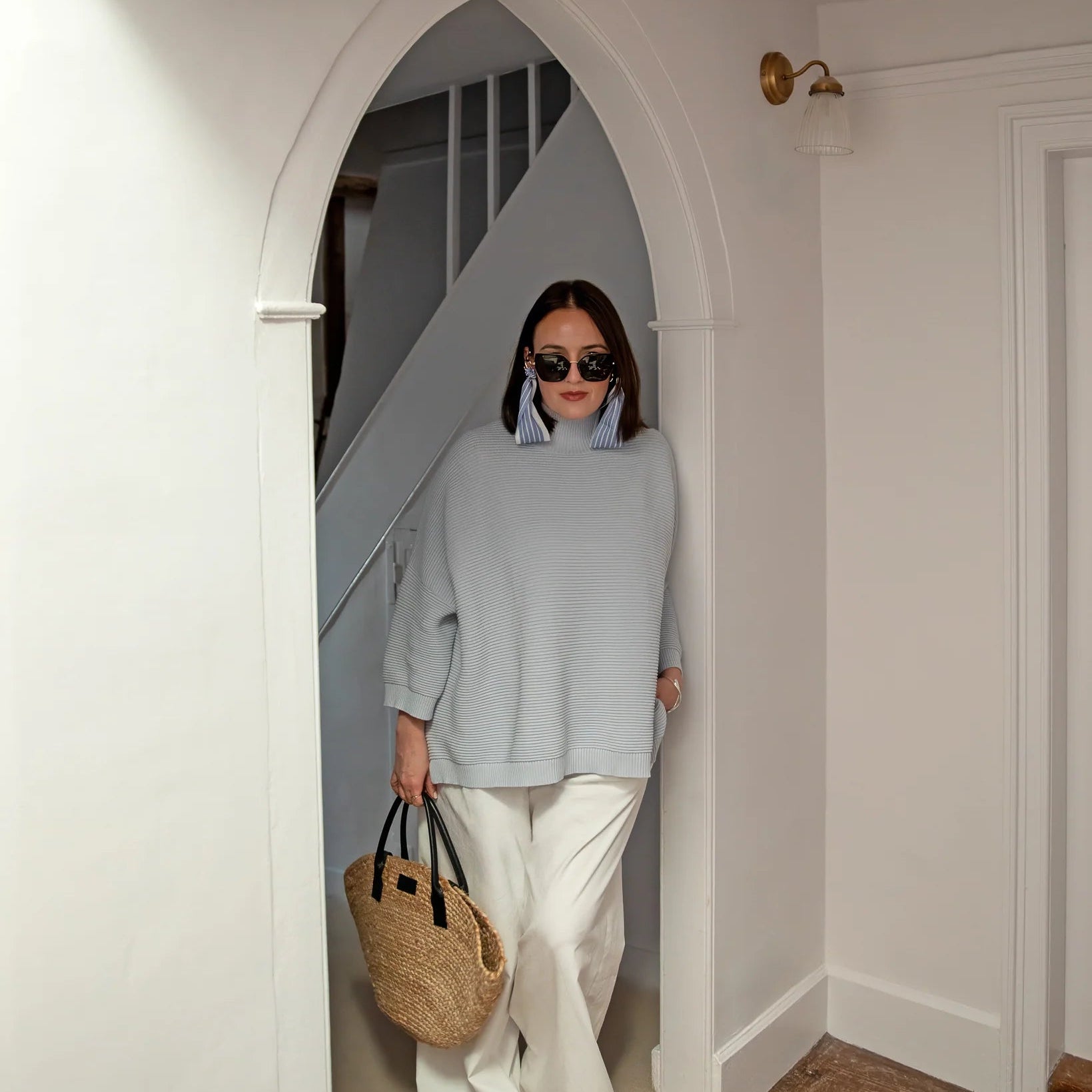 Woman in a light blue sweater and white pants standing in a doorway holding a straw bag.
