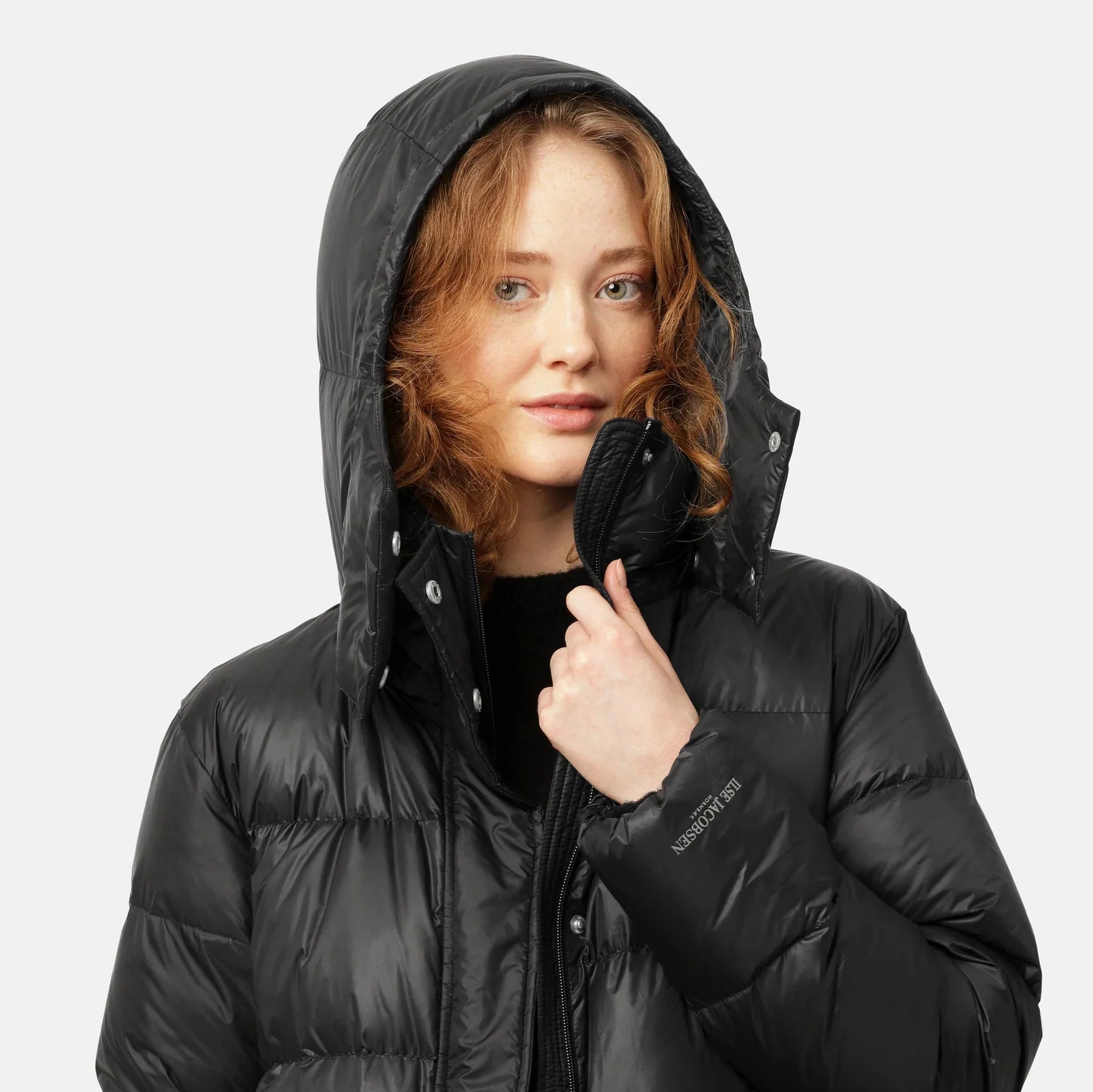 Black Lise Jacobsen Honnørx puffer jacket with a hood, worn by a woman with reddish-brown hair. The jacket has a shiny, quilted
