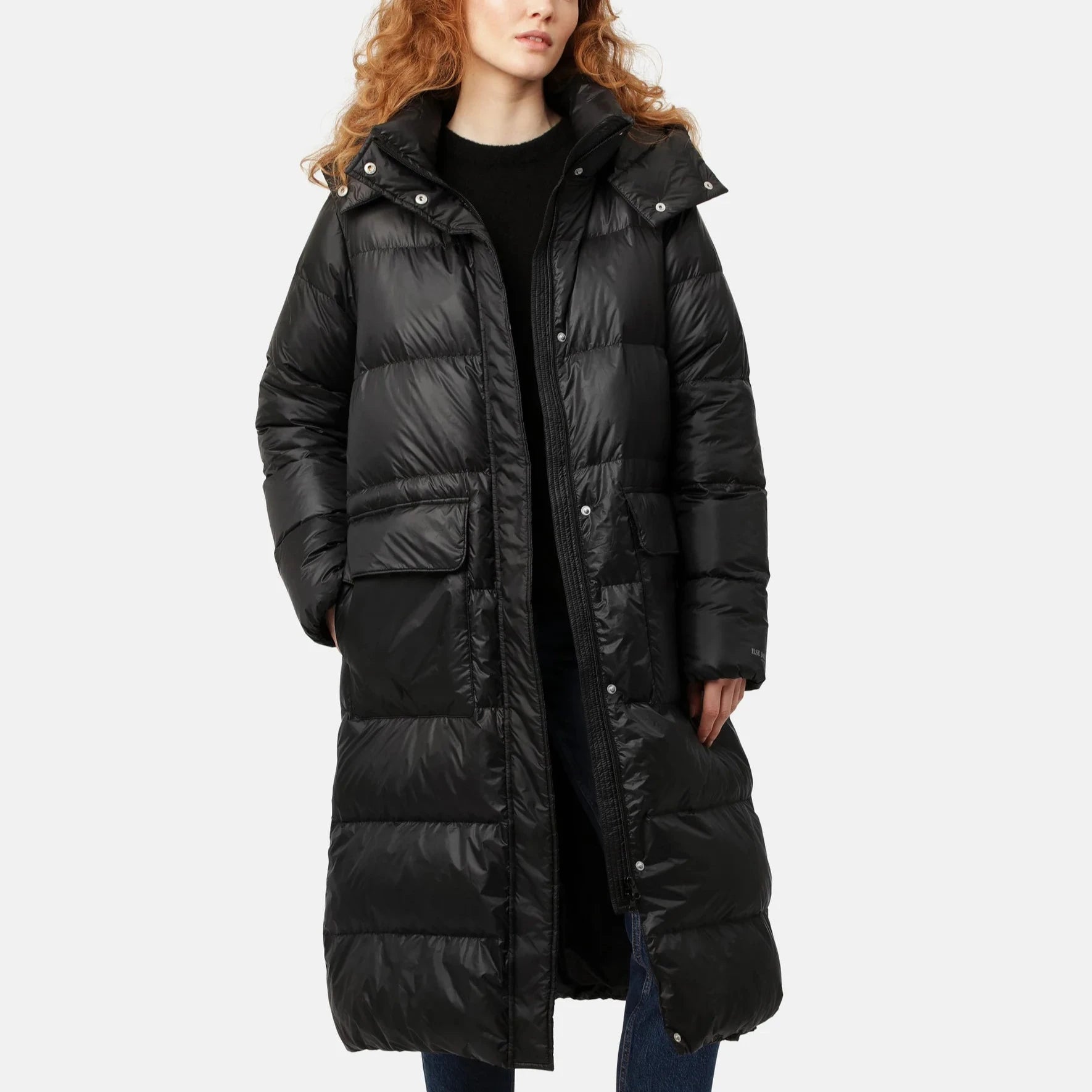 Black long puffer coat with a hood, featuring a shiny, quilted texture and snap closures. The model wears jeans underneath.