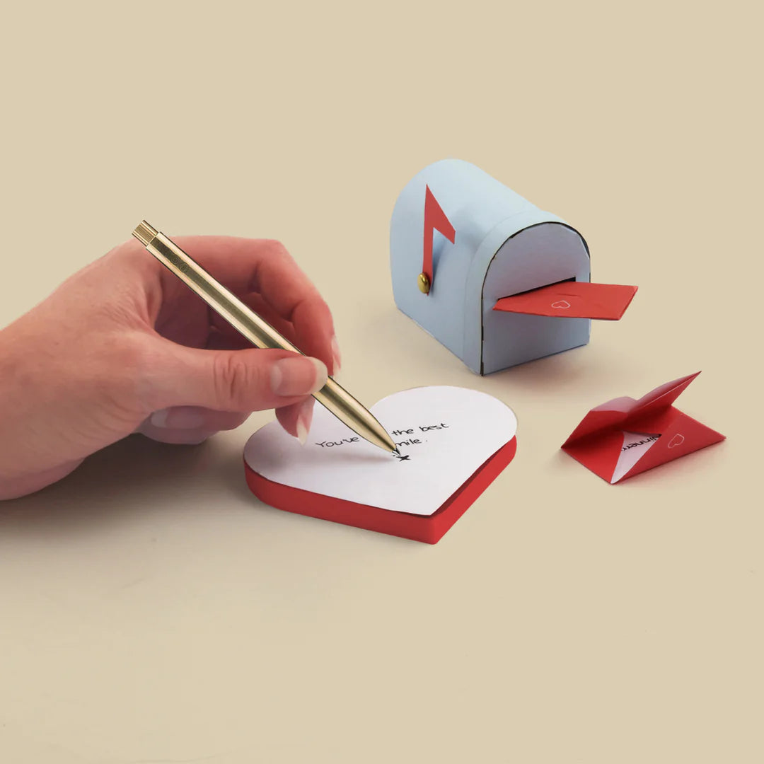 Heart-shaped notepad with a pen, next to a small mailbox on a beige background