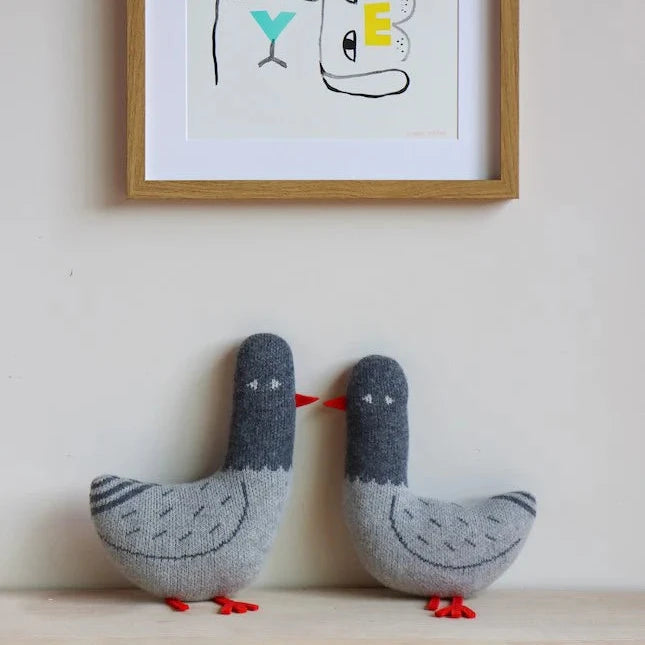 Two soft, knitted gray pigeons with red beaks and feet by Donna Wilson, displayed beside a colorful framed artwork.