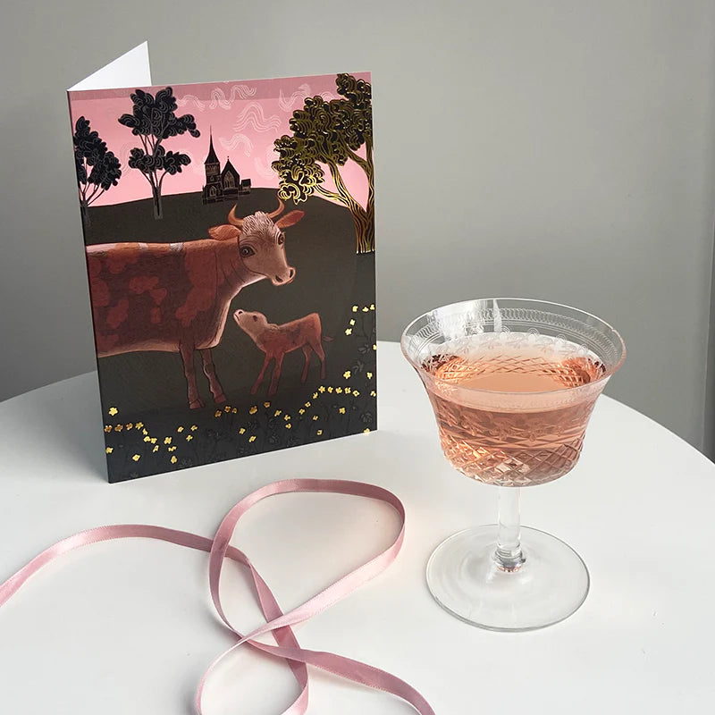 Greeting card with cow illustration next to a glass of pink cocktail on a white surface.