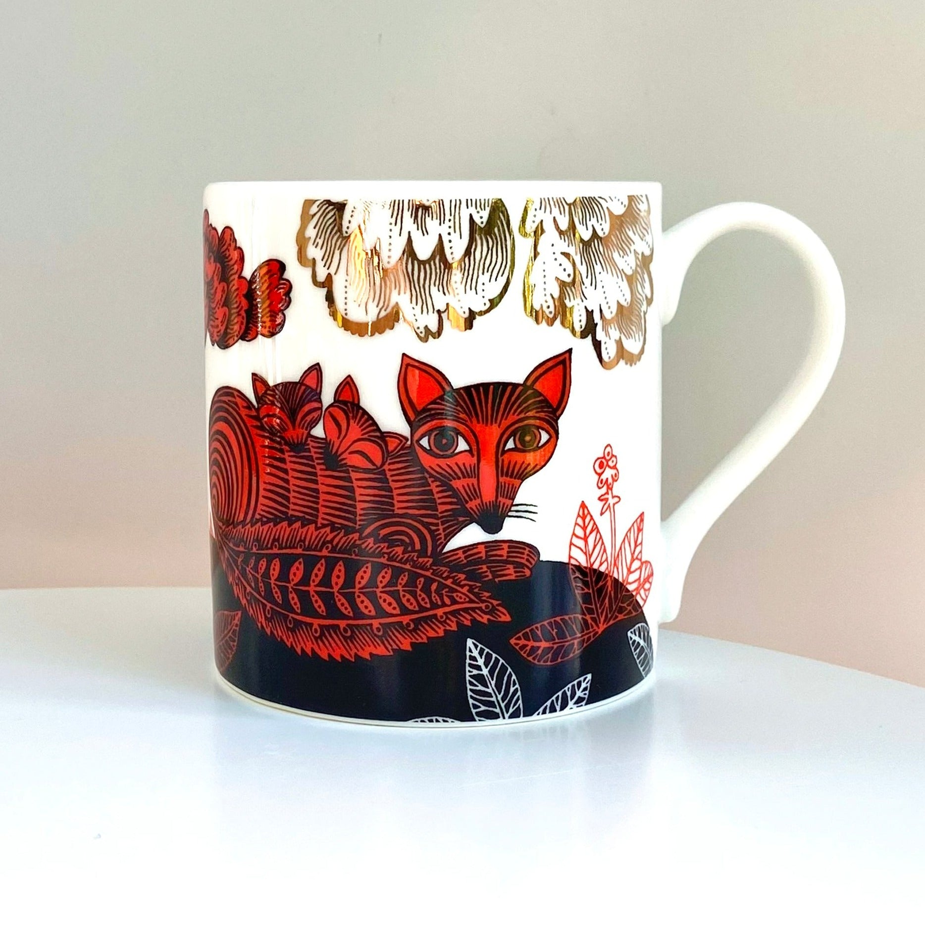 White mug with a red and black illustration of a fox and kits nestled amongst stylized foliage and gold floral accents.