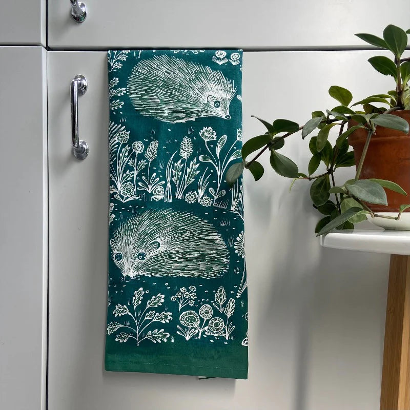Dark teal tea towel featuring a white line drawing of two hedgehogs amidst wildflowers.