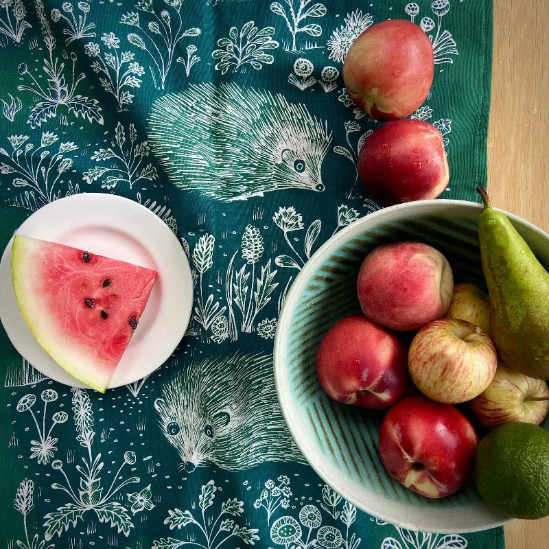 Green tablecloth with hedgehog print, featuring a bowl of fruit (peaches, apples, pear) and a slice of watermelon on a plate.