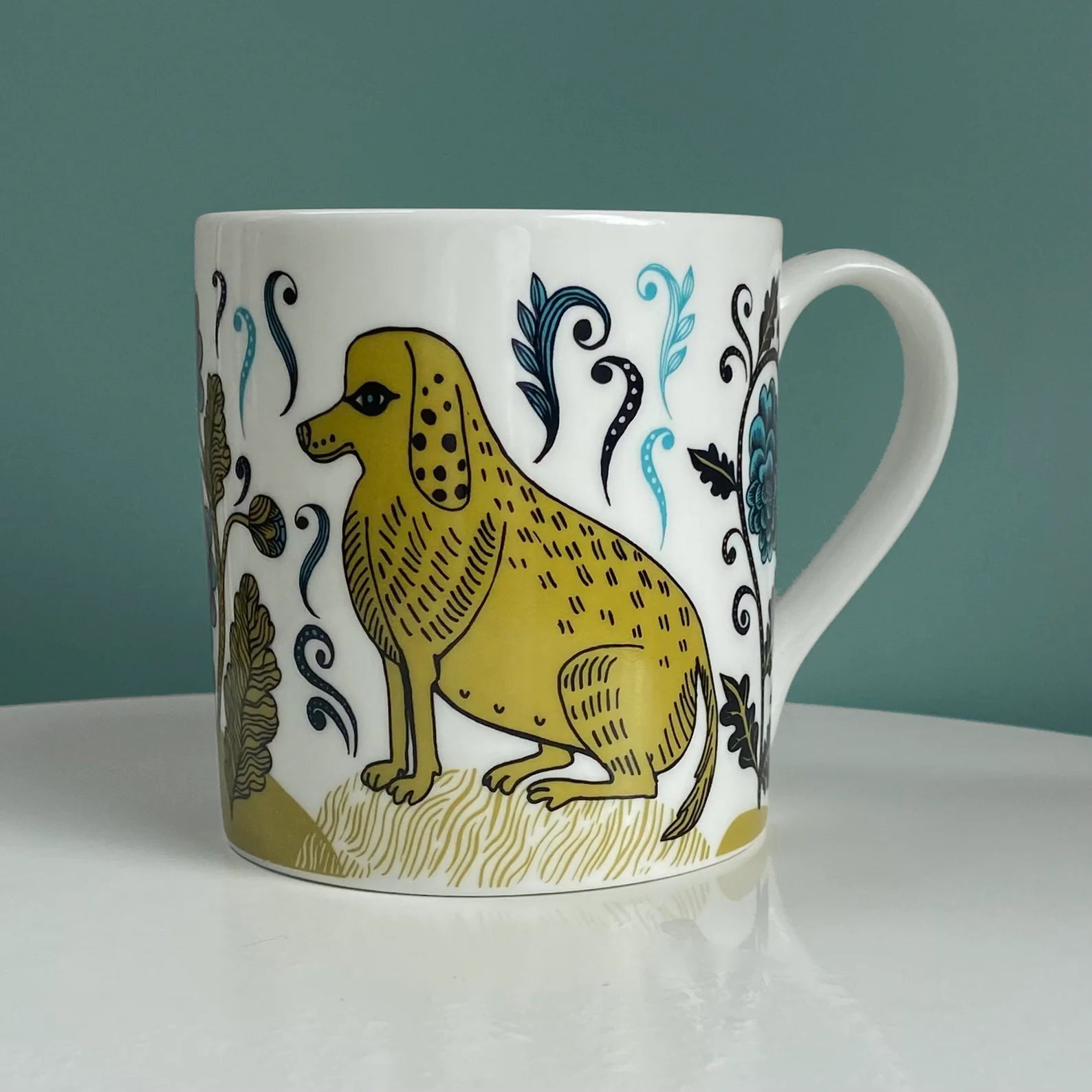 White mug featuring a mustard-yellow dog illustration with black detailing, surrounded by stylized teal and gold floral