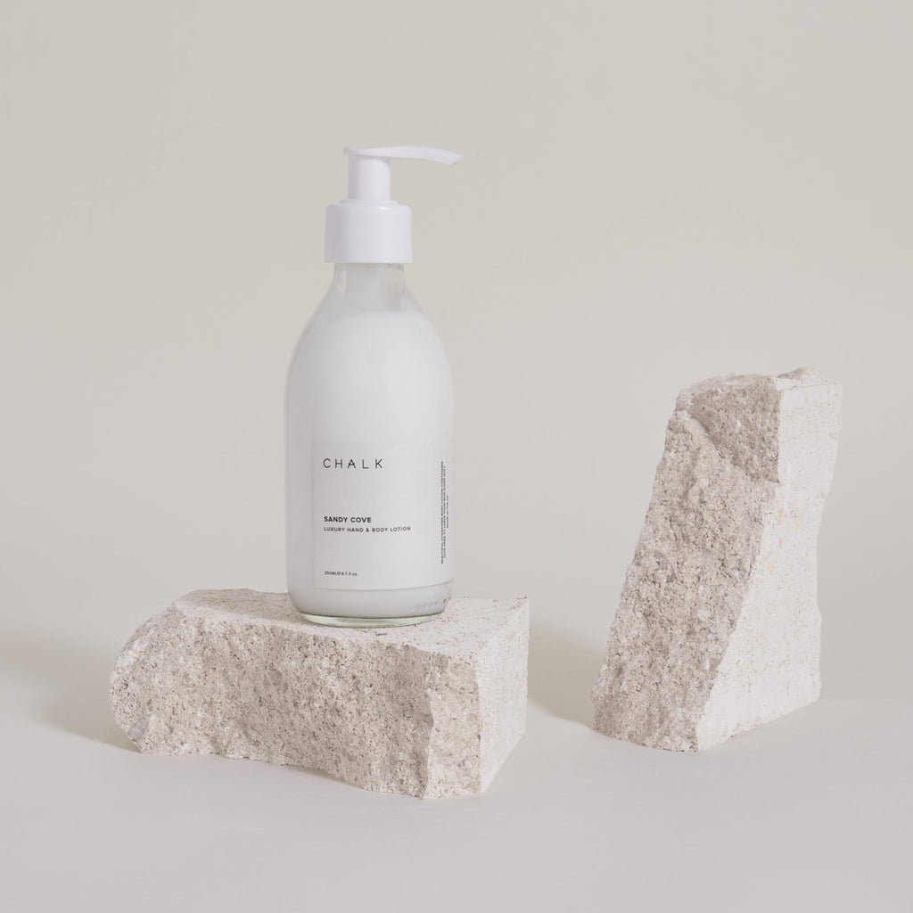 A bottle of CHALK Sandy Cove luxury hand and body lotion on textured beige stones against a light background.