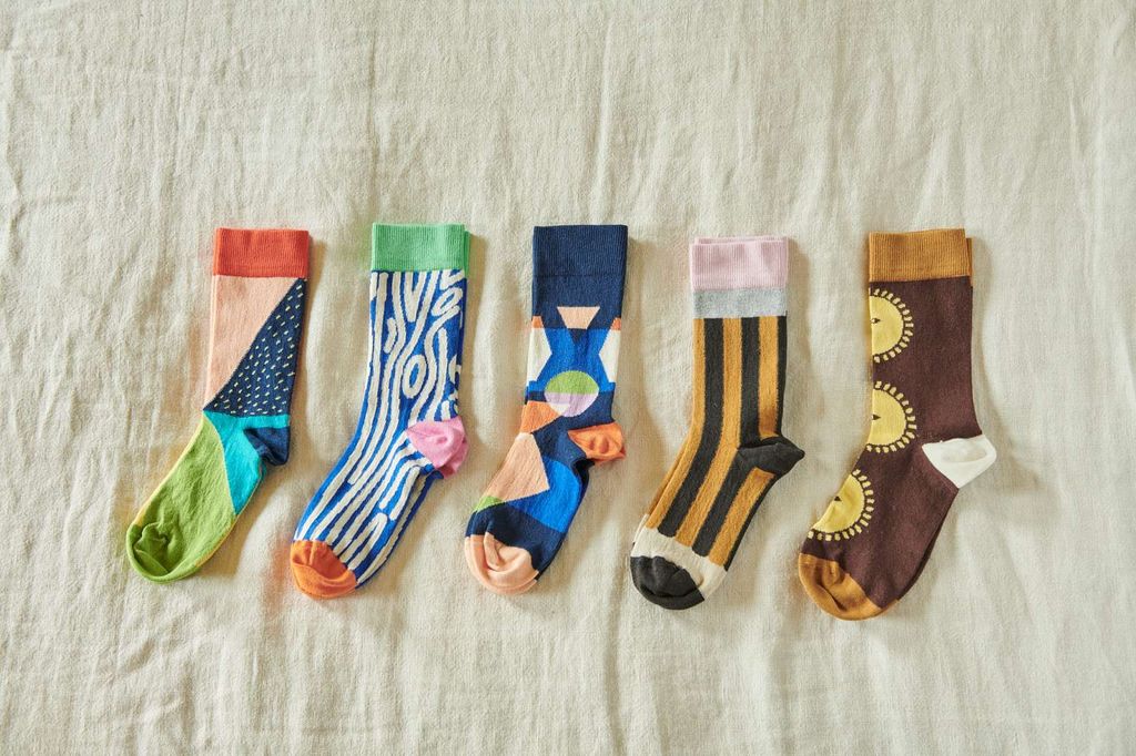 Five pairs of colorful patterned socks laid flat on a light beige fabric background. The socks feature various designs including stripes, wavy lines, sun motifs, and abstract shapes in blue, green, orange, pink, yellow, and brown.