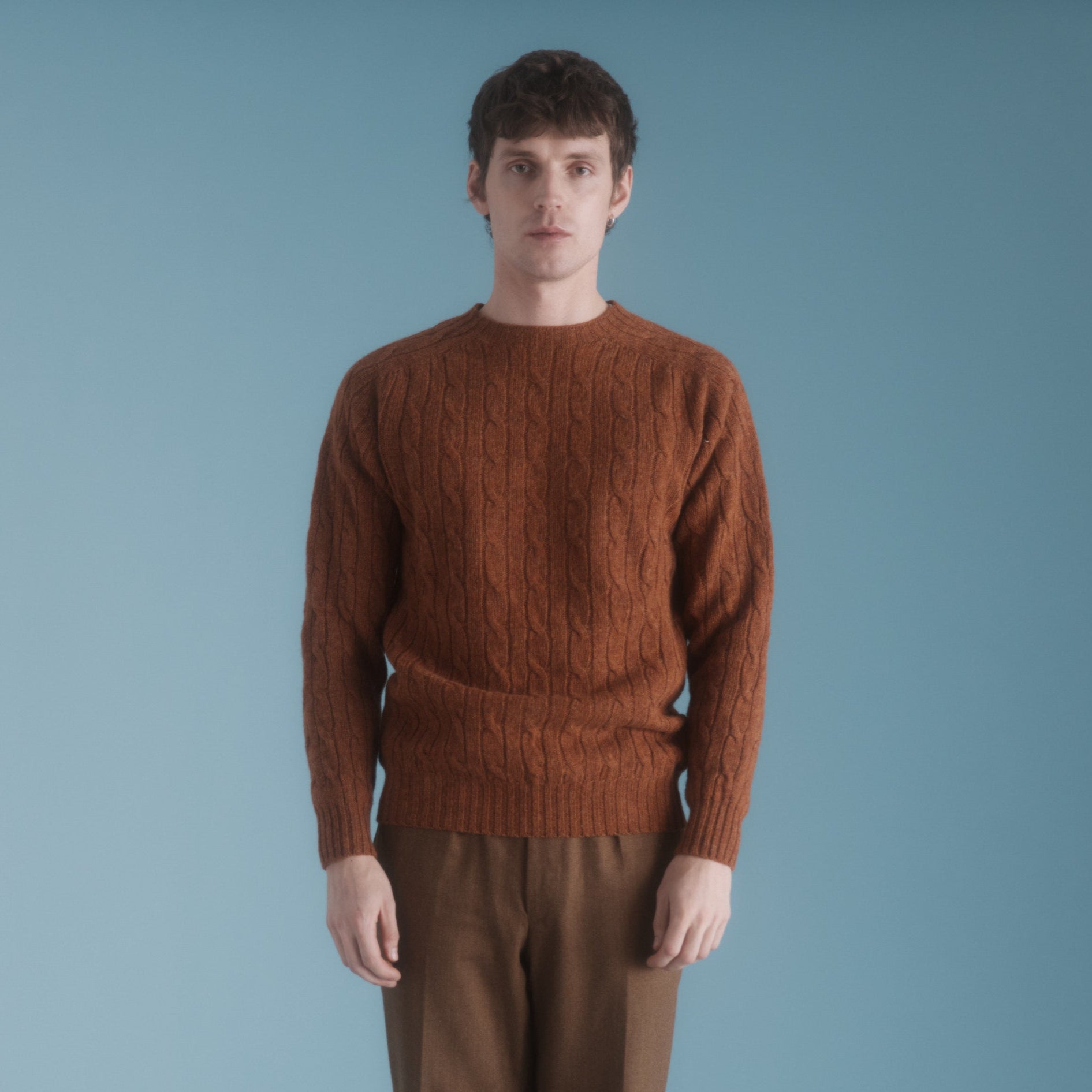 A rust-colored cable-knit wool sweater. The sweater is shown on a model against a light blue background.