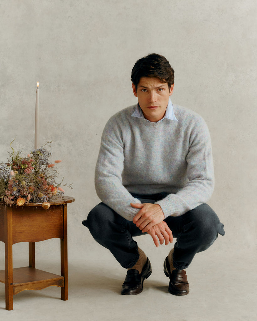a male model crouching by a wooden table with dried flowers and a lit candle, wearing a blue collared shirt and a Harley of Scotland Men's Jumper - Ugie Pearl