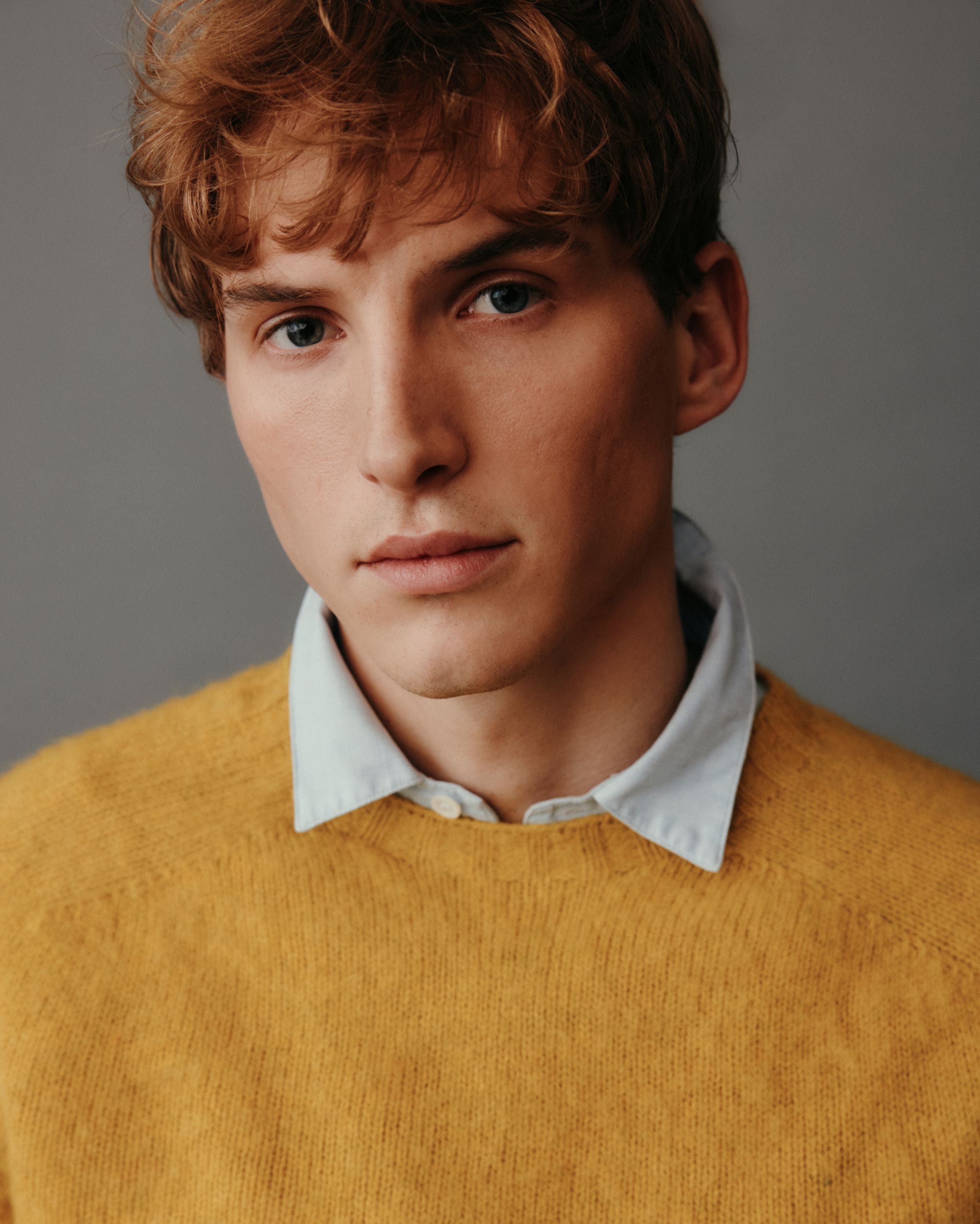 Man wearing a mustard yellow sweater over a white shirt against a gray background