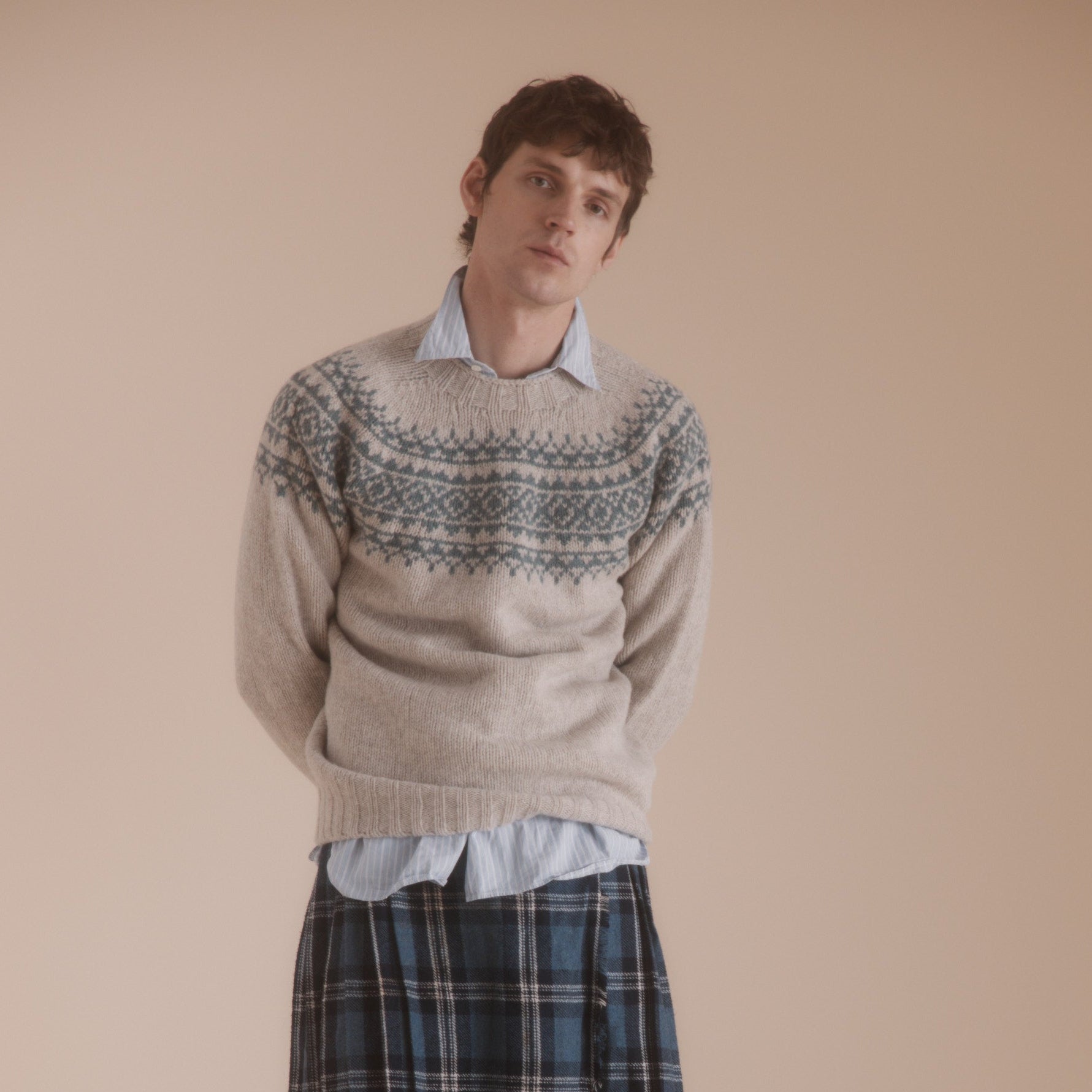 Beige crewneck sweater with teal Fair Isle pattern, worn over a light blue shirt and a blue, black, and white plaid kilt.