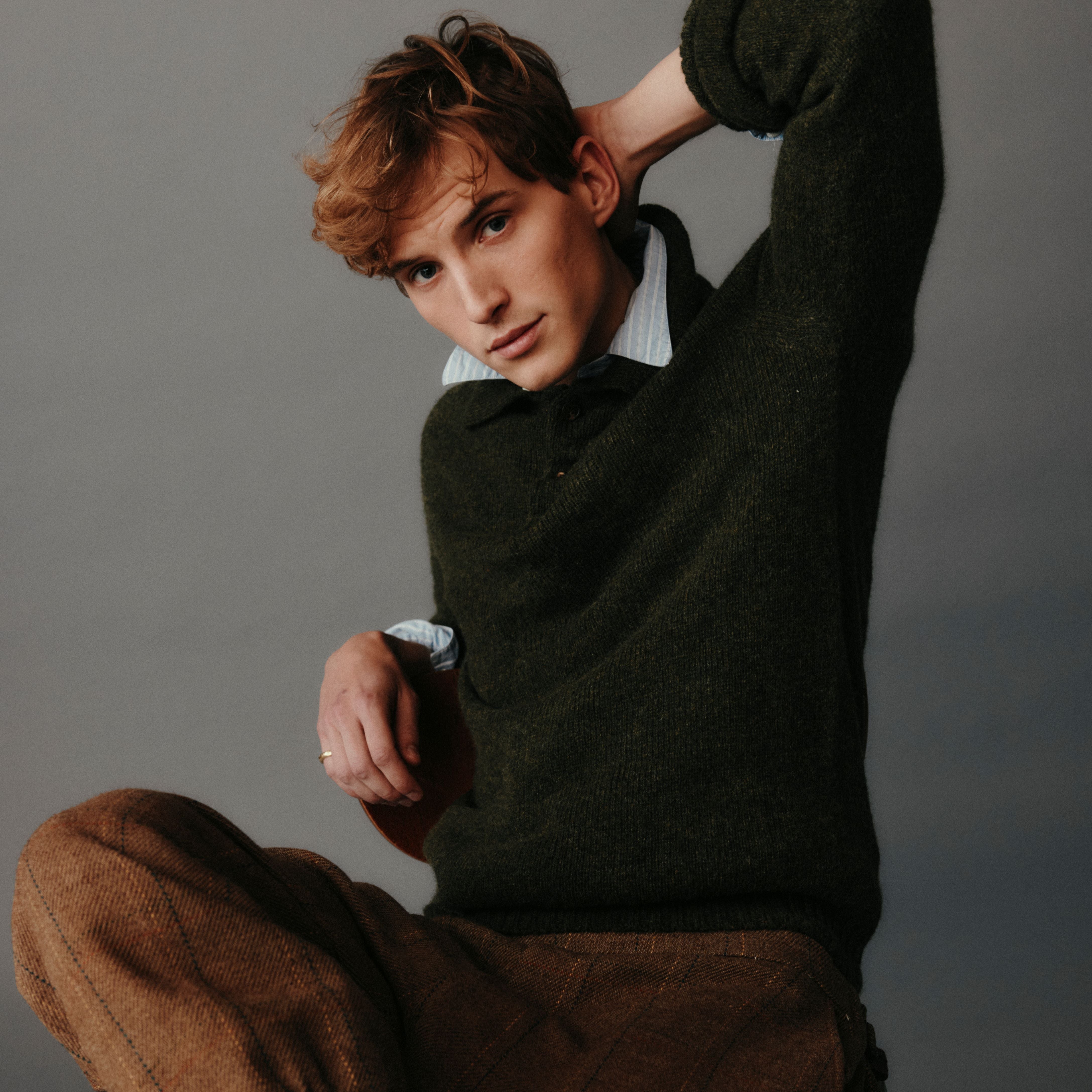 Man wearing a dark green sweater and brown pants against a gray background