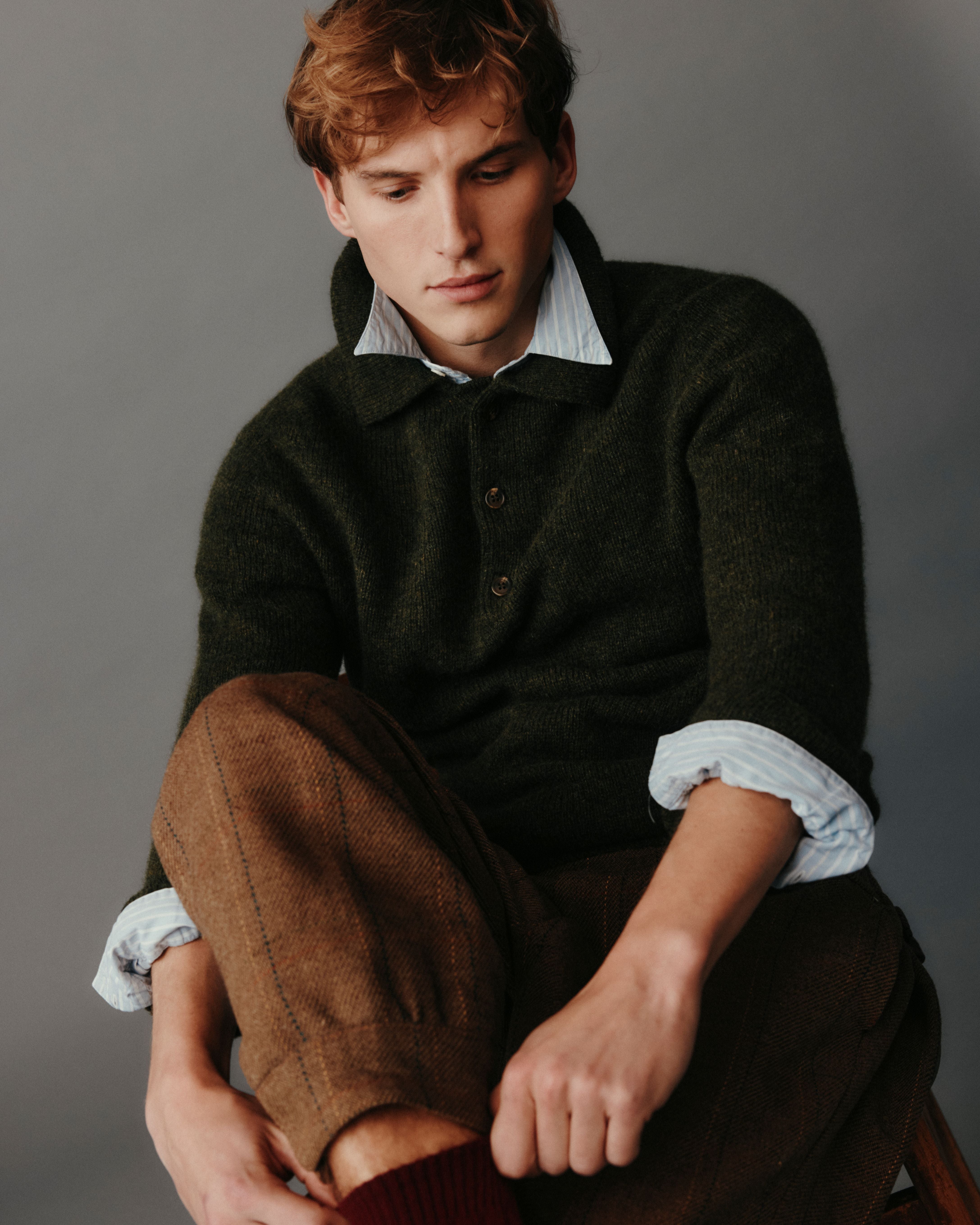 Man sitting on a stool wearing a dark sweater and brown pants against a gray background