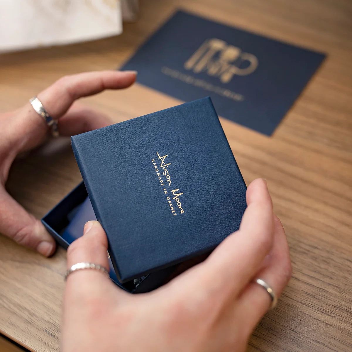 Person opening a blue jewelry box with a brand logo on a wooden surface