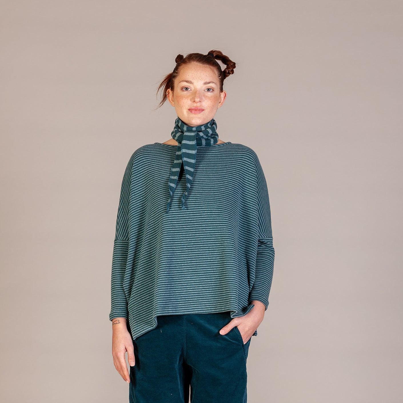 A woman stands against a plain beige studio background wearing a teal-green striped long-sleeve poncho top with a matching striped neck scarf, dark teal pants, and brown shoes.