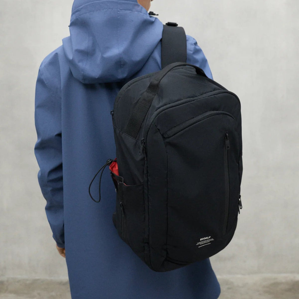 A person in a blue Ecoalf jacket carries a black backpack with red accents on the side.