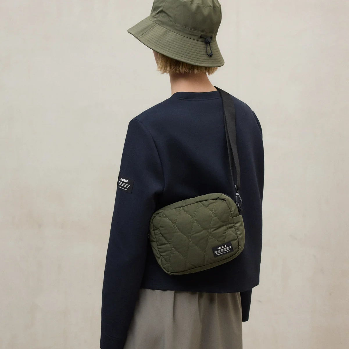 Person wearing a navy sweater, beige pants, and a green bucket hat with a green crossbody bag against a plain background.
