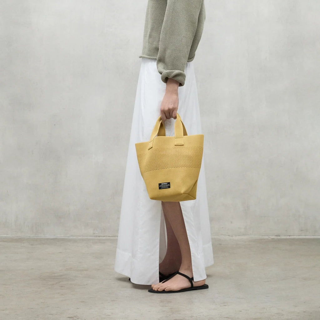 Person wearing a light green sweater and a white skirt, holding a yellow Ecoalf bag.