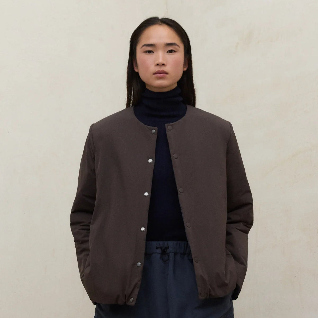 Person wearing a brown jacket over a dark turtleneck and pants on a beige background