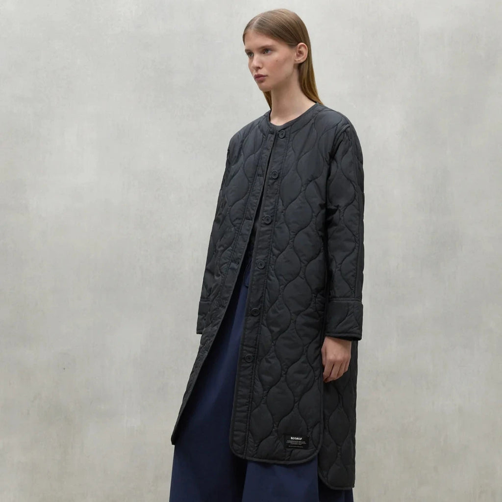 A model in a black Ecoalf quilted coat and navy blue pants stands against a textured gray background.