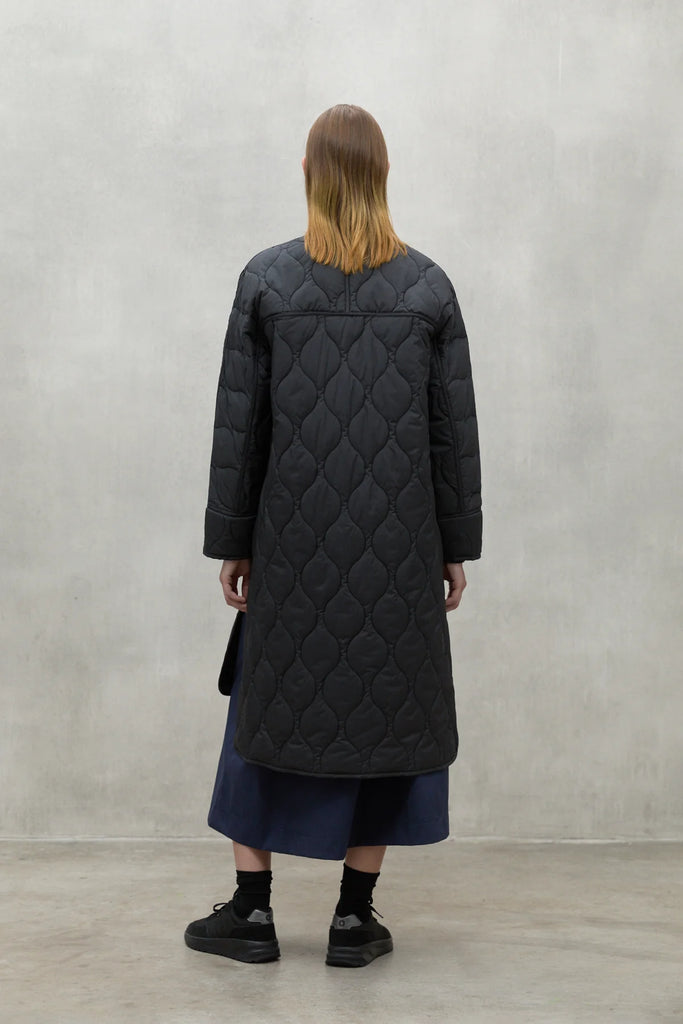 A model stands with their back to the camera, wearing a black Ecoalf quilted coat and a dark blue skirt.