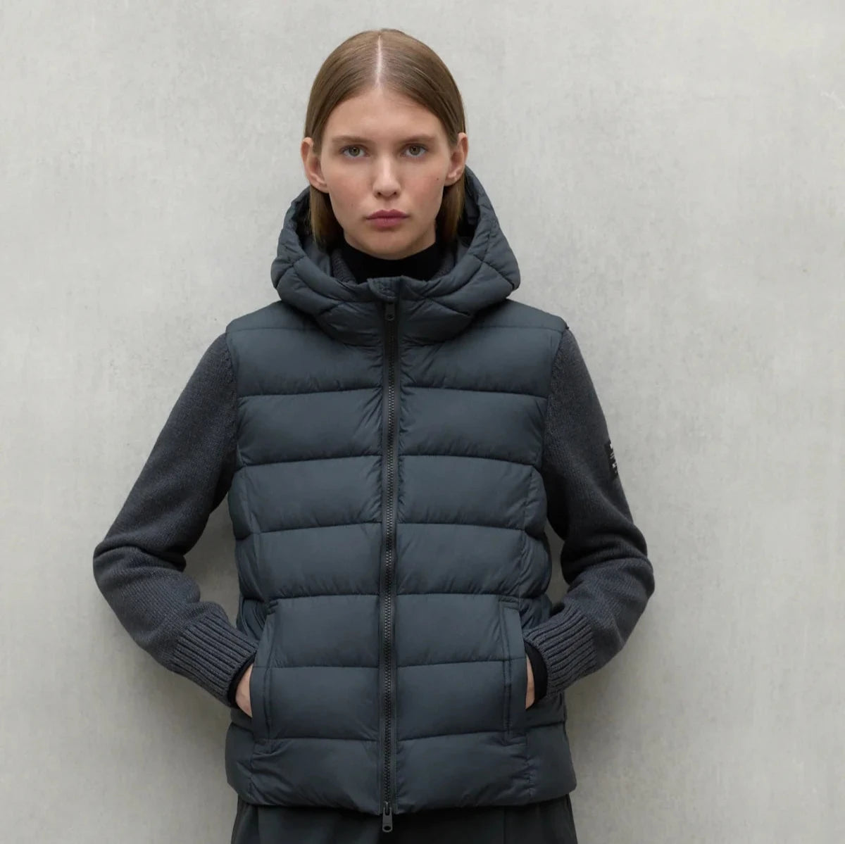 Dark teal hooded puffer vest with gray knit sleeves, zipper closure, and front pockets.