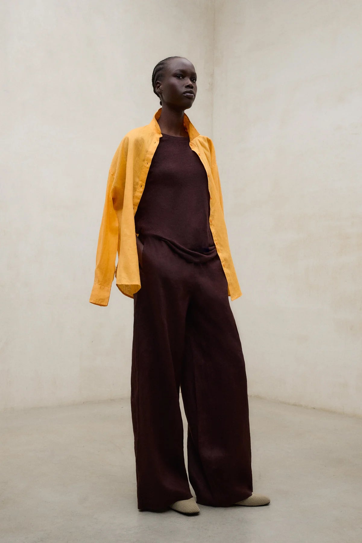 Person wearing a mustard yellow jacket over a brown top and pants on a plain background