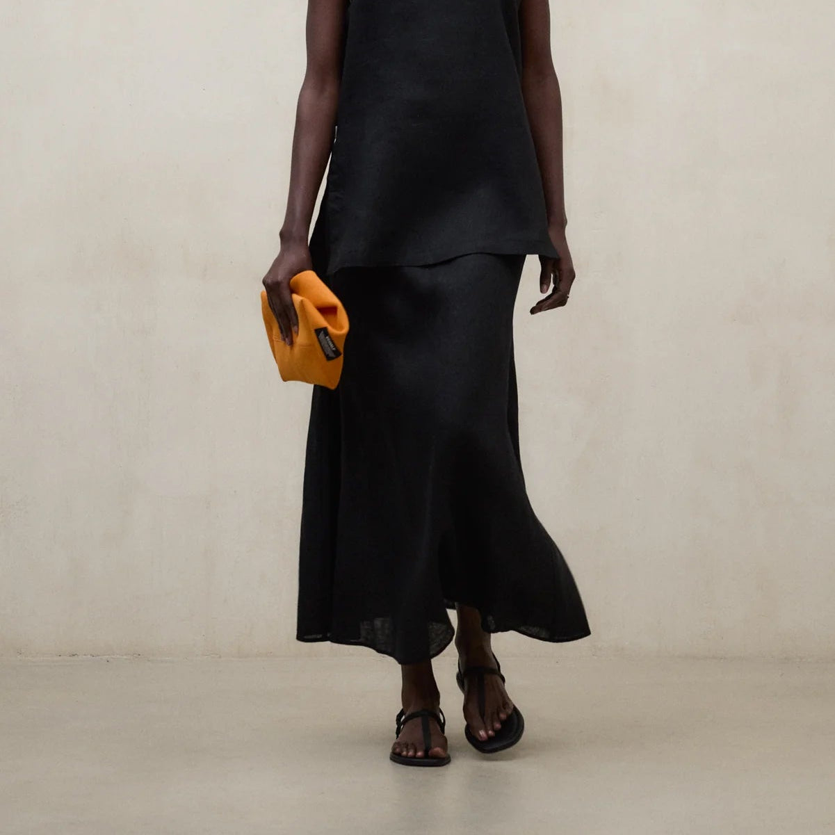 Woman wearing a black sleeveless top and skirt holding a yellow clutch against a beige background