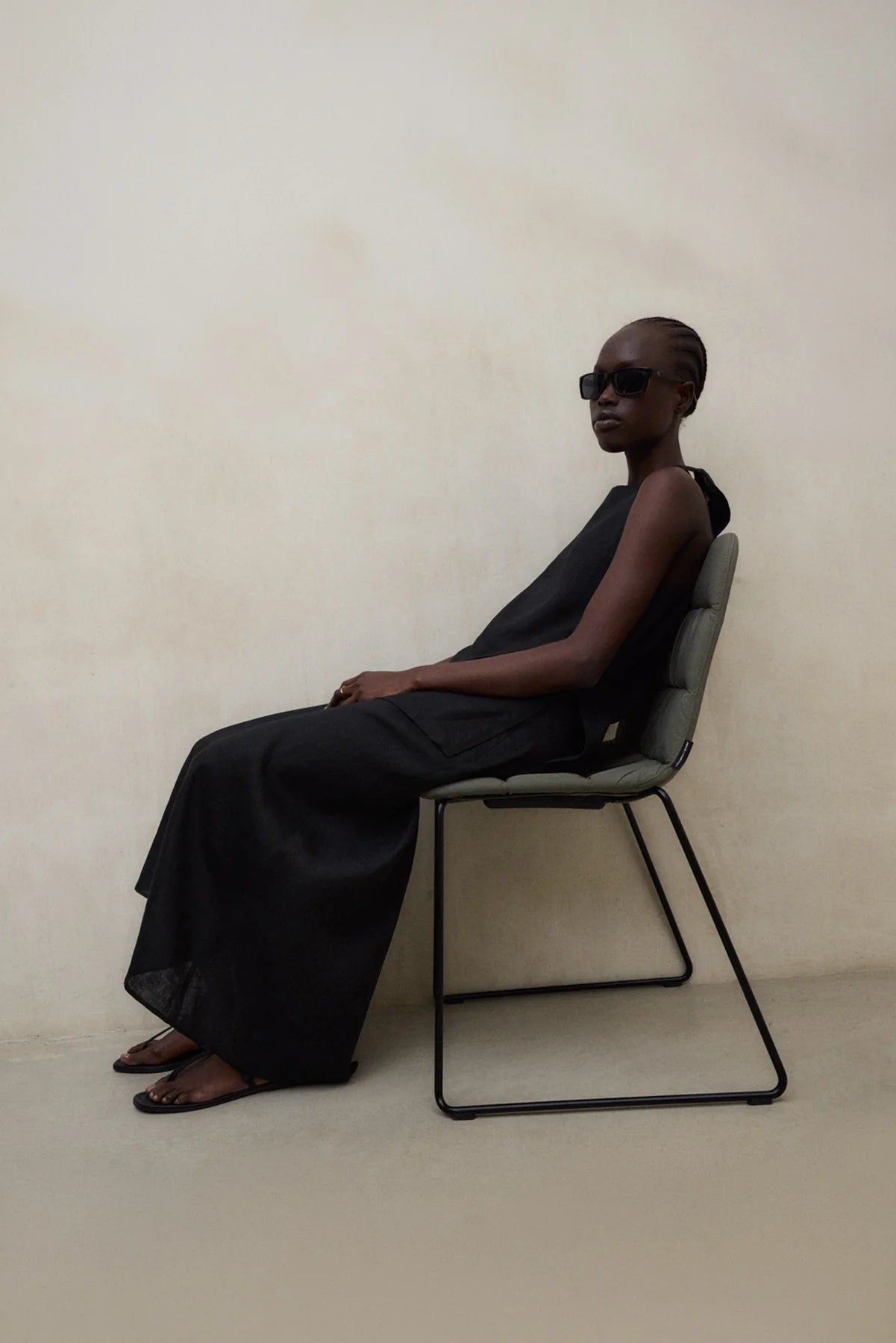 Person wearing a black outfit sitting on a chair against a plain background