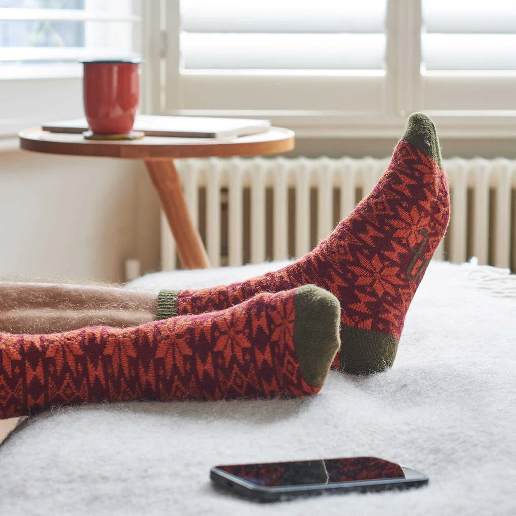 Red patterned socks with green toes and cuffs on a bed, with a phone and cup in the background.