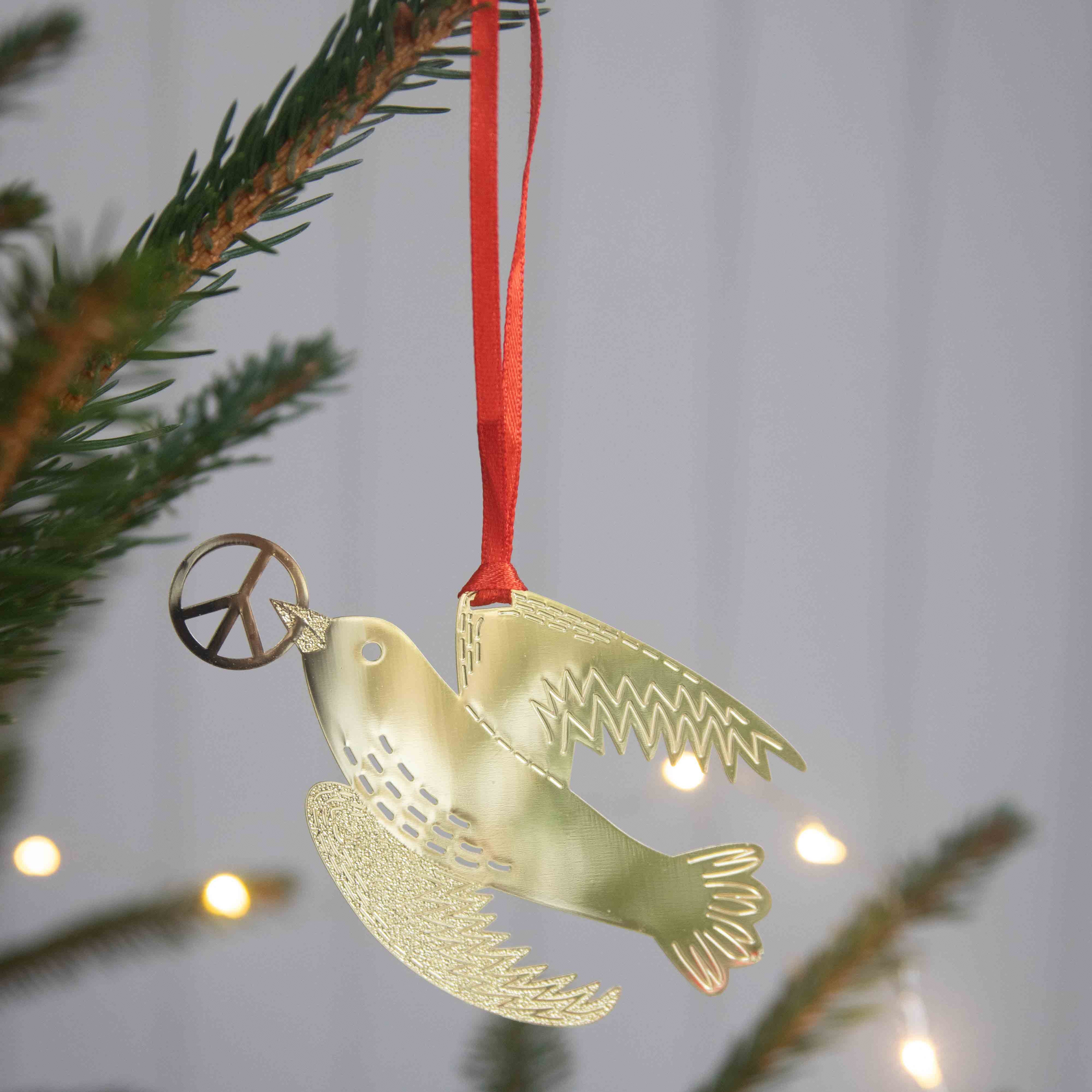 A metallic gold dove Christmas ornament holding a peace sign, hanging from a red ribbon on a pine tree branch with warm string lights in the background.