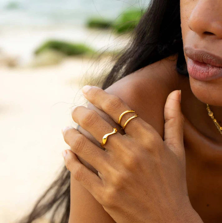 A close-up lifestyle shot of a woman on a beach wearing two gold rings on her hand, with the ocean and sand blurred in the background.