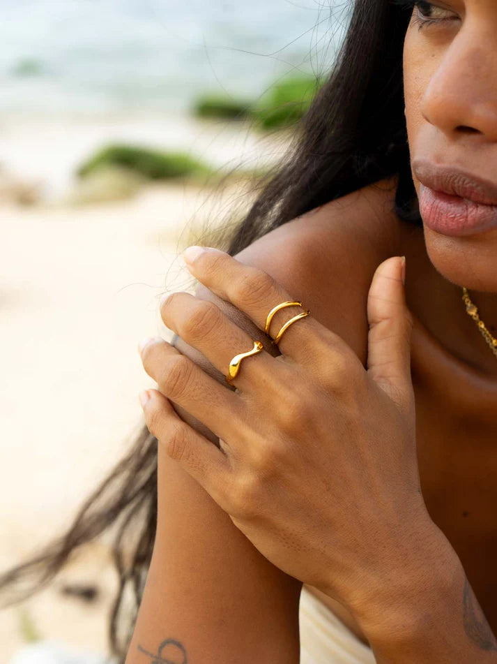 A close-up lifestyle shot of a woman on a beach wearing two gold rings on her hand, with the ocean and sand blurred in the background.