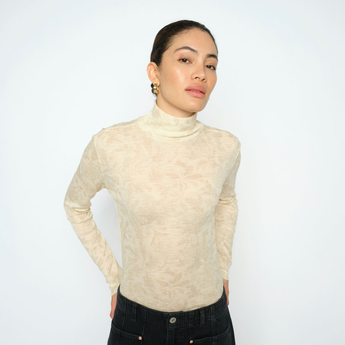 Woman wearing a beige turtleneck sweater and black pants against a white background