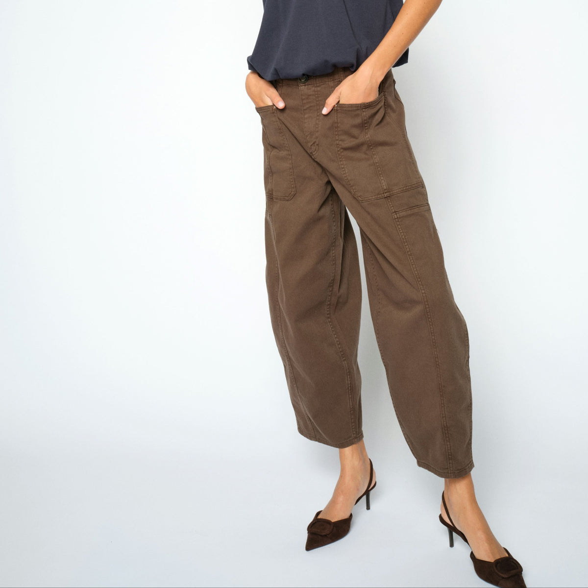 A woman modeling a loose navy blue short-sleeve top and brown wide-leg cropped pants with dark brown pointed-toe heels, standing against a light gray studio background.