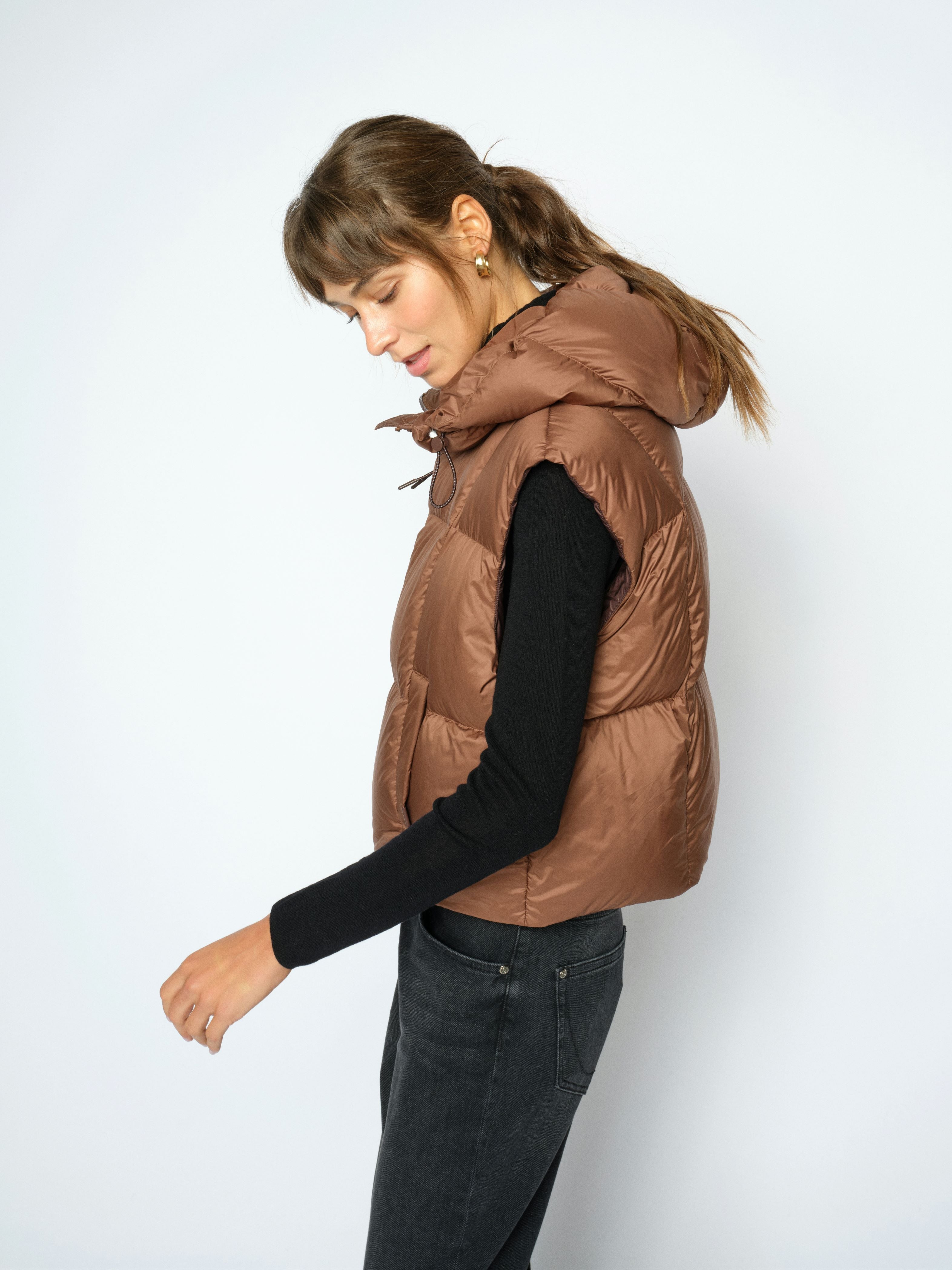 A woman wearing a brown puffer vest over a black long-sleeve top and dark jeans, photographed against a plain light gray studio background.