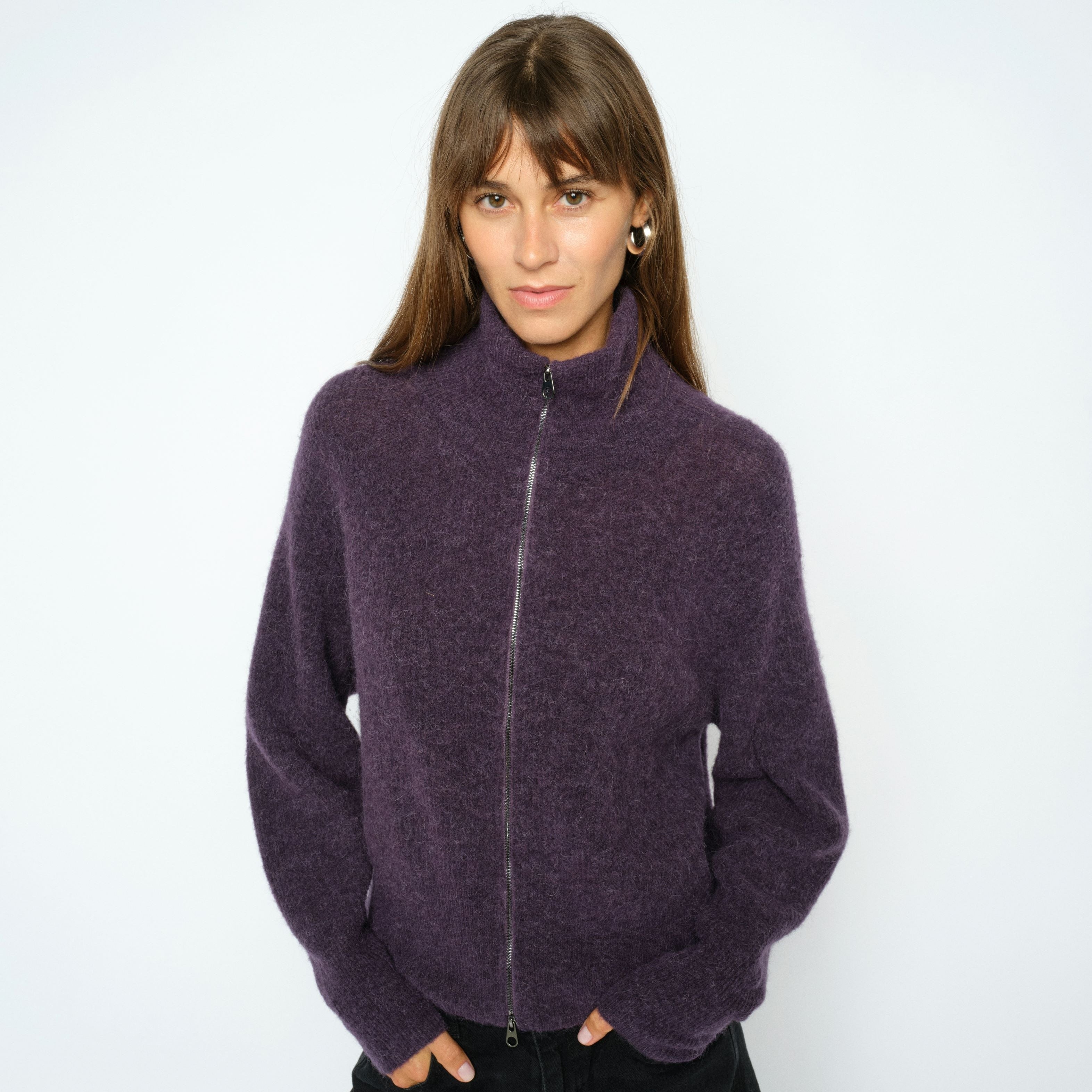 A woman modeling a fuzzy, short-sleeved purple zip-up fleece jacket with a mock neck, paired with black pants, against a plain light gray background.