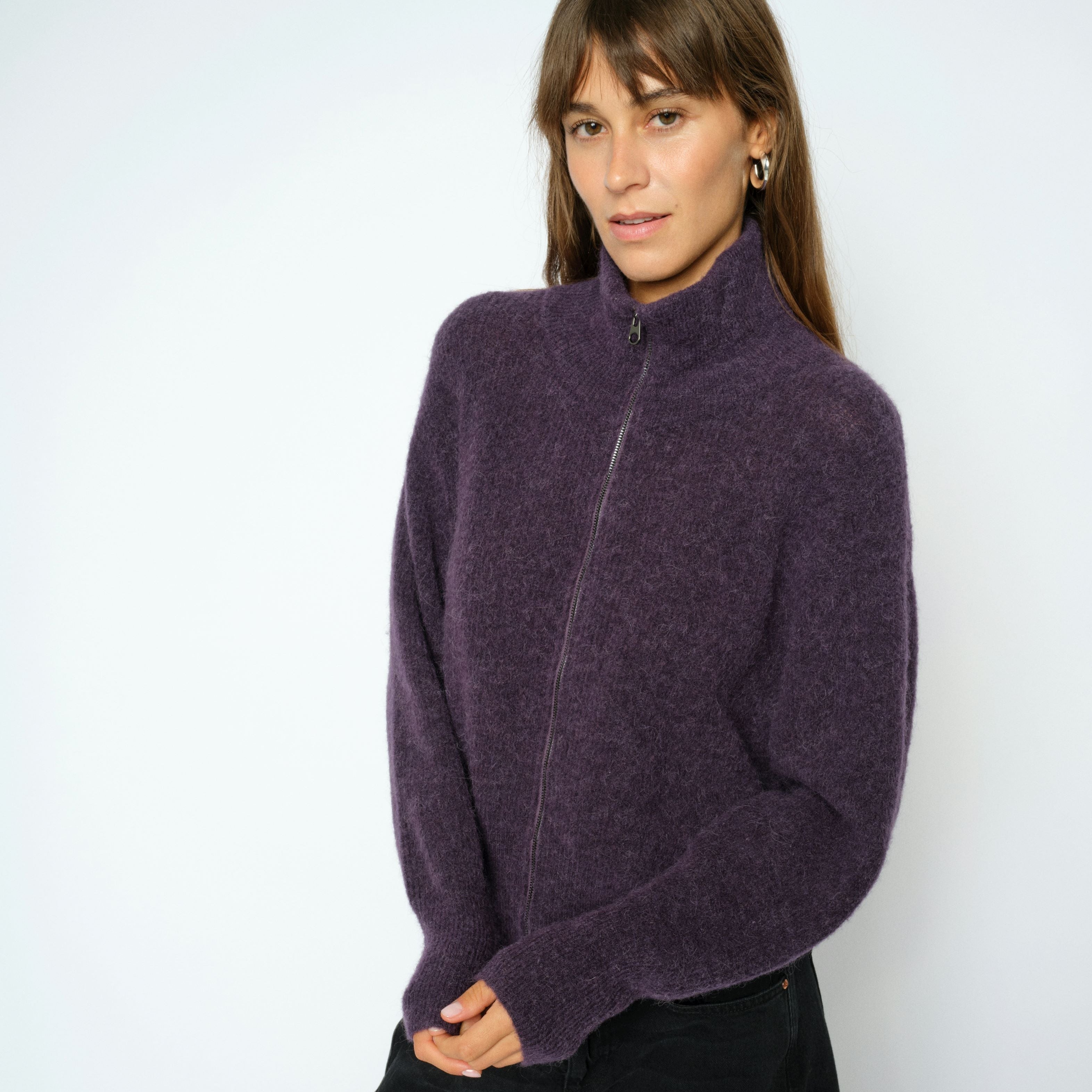 A woman wearing a fuzzy purple mock-neck zip-up sweater and black pants, standing against a plain light gray studio background.