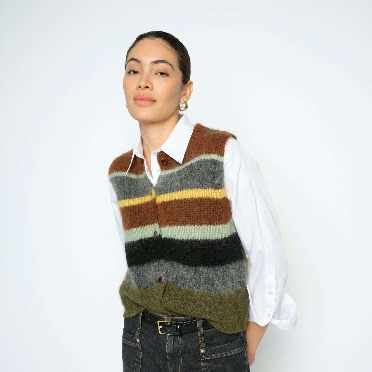 A model wearing a brown, beige, black, and olive green striped knit sweater vest over a white collared shirt and dark gray jeans, standing against a plain white studio background.