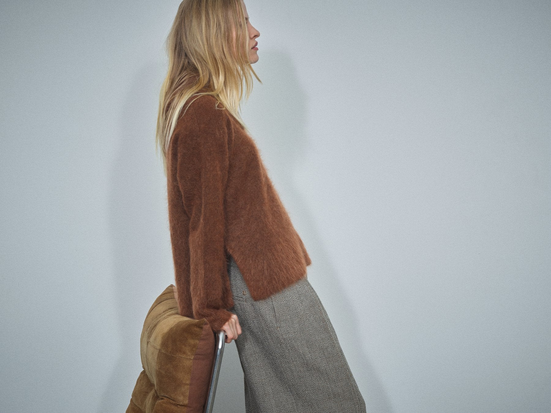 A woman in profile wearing a fuzzy brown cropped cardigan and a gray skirt, holding a brown leather bag against a plain light gray wall.