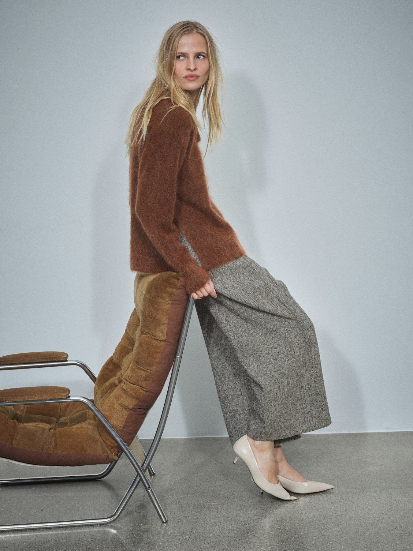 A woman models a brown fuzzy knit sweater and wide-leg gray trousers, leaning against a modern brown chair in a minimalist studio setting.