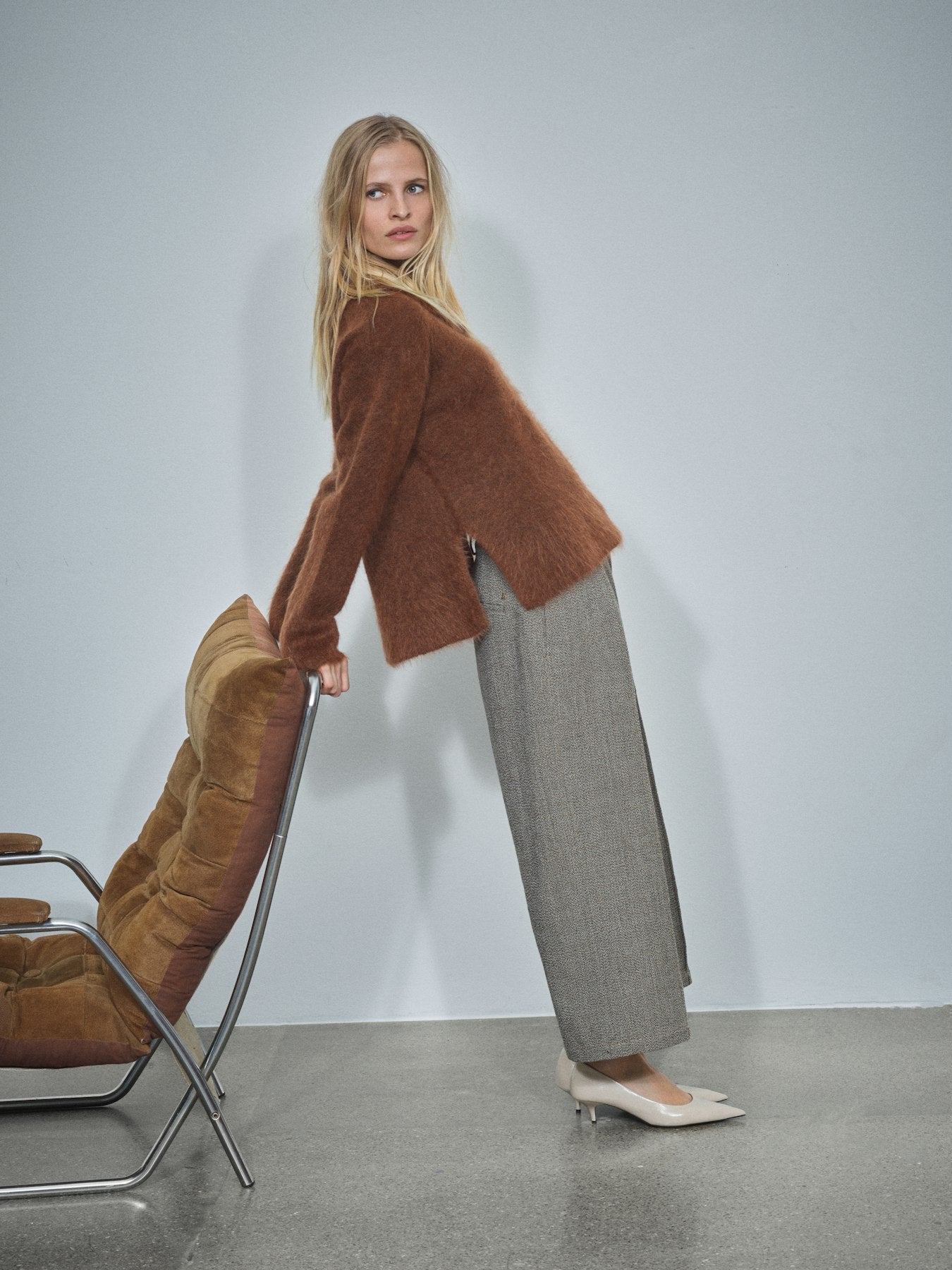 A woman models a brown fuzzy cropped cardigan and wide-leg gray trousers, standing next to a modern brown chair against a light gray studio wall.