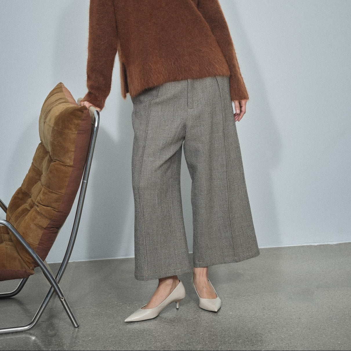 A woman stands in a studio wearing a fuzzy brown long-sleeve top and wide-leg gray trousers with white pointed-toe heels, posed beside a brown upholstered chair against a light gray wall.