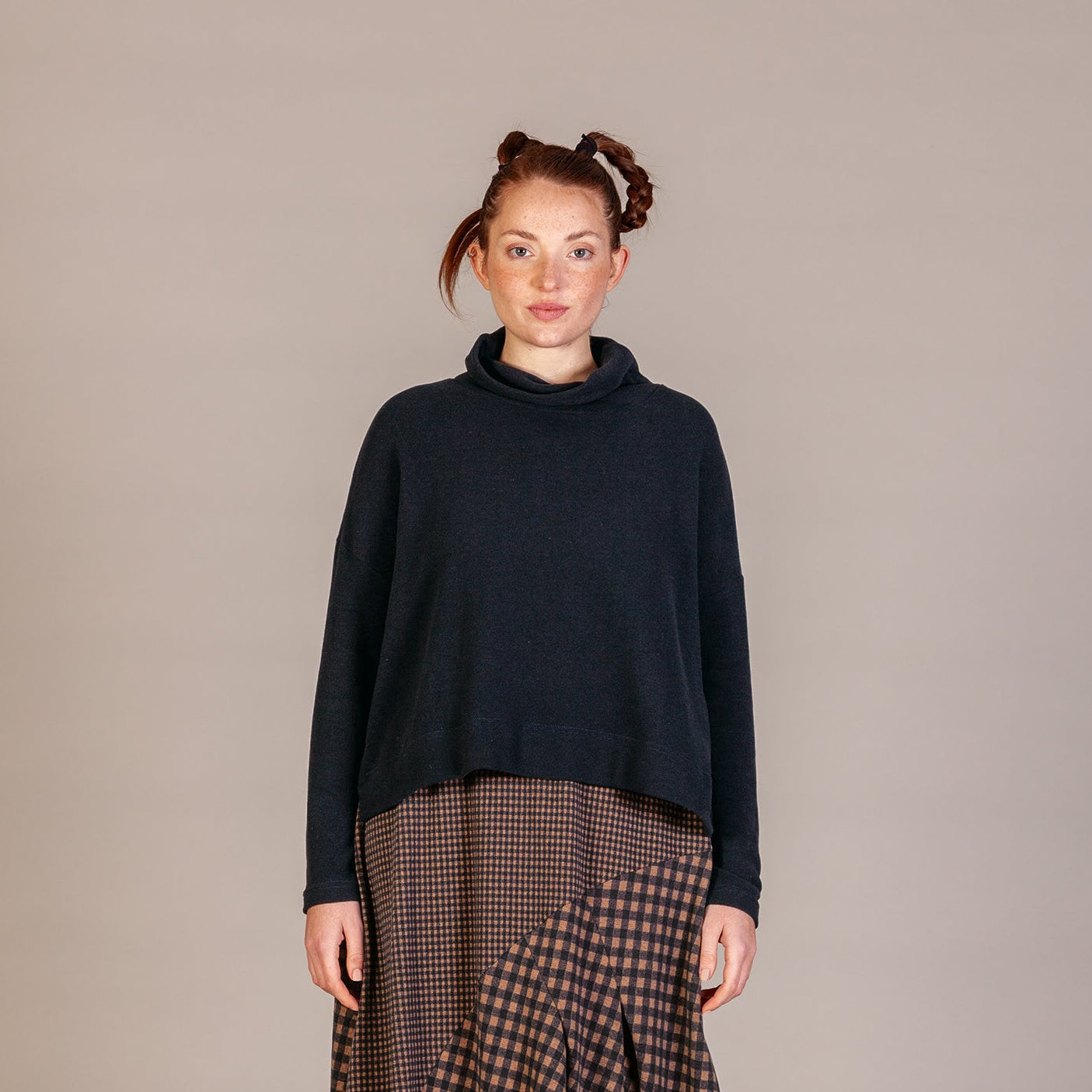 A model stands against a neutral studio backdrop wearing a dark navy turtleneck sweater and a brown and black patterned midi skirt with lace-up boots.