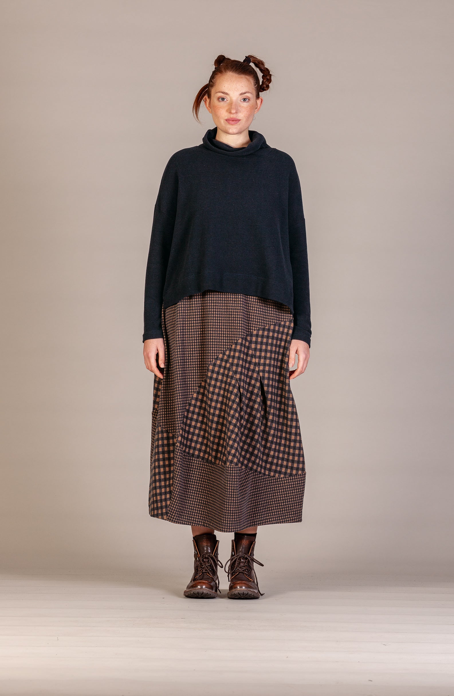 A model stands against a neutral studio backdrop wearing a dark navy turtleneck sweater and a brown and black patterned midi skirt with lace-up boots.
