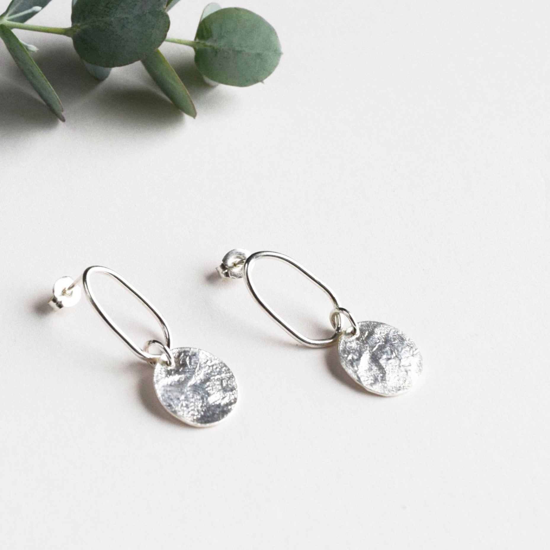 Silver earrings with textured circular pendants hanging from oval wire loops.